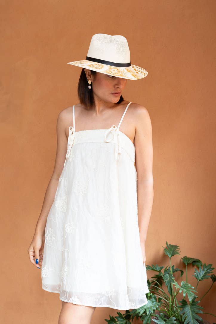 Woman wearing a white dress and an artisan painted gold and white hat against a brown background. Crafted in Oaxaca, Mexico.