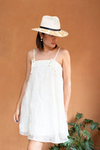 Woman wearing a white dress and an artisan painted gold and white hat against a brown background. Crafted in Oaxaca, Mexico.