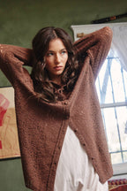 A woman with long wavy brown hair wearing a textured brown button-up cardigan over a white dress, posing with her arms raised in an indoor setting.