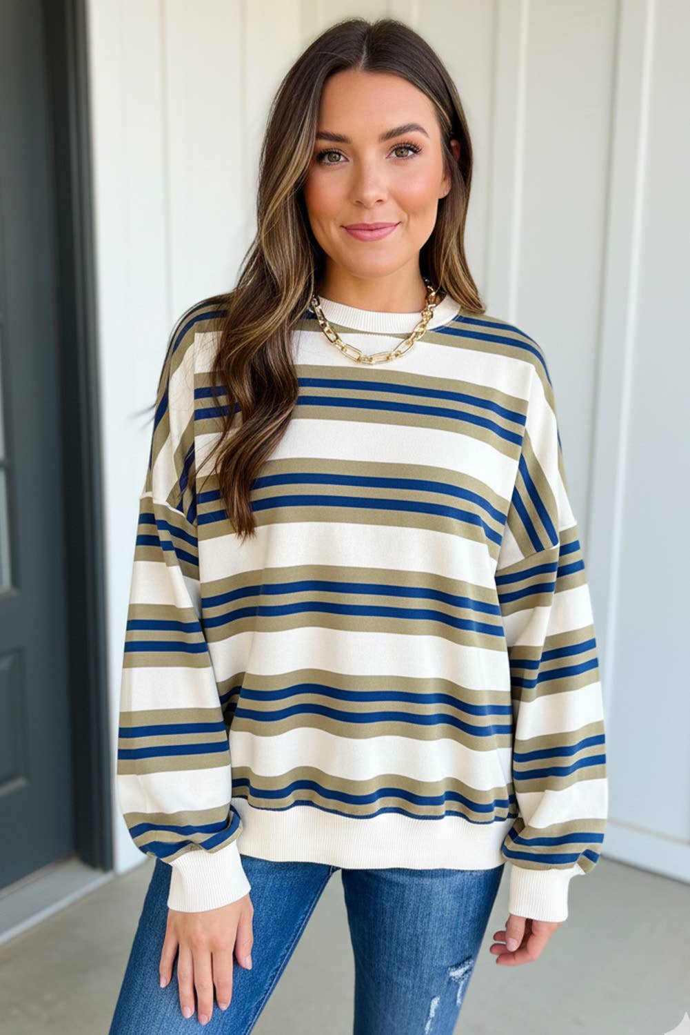 A woman wearing a cream-colored knit sweater with horizontal navy blue and olive green stripes, paired with blue jeans and a gold chain necklace.