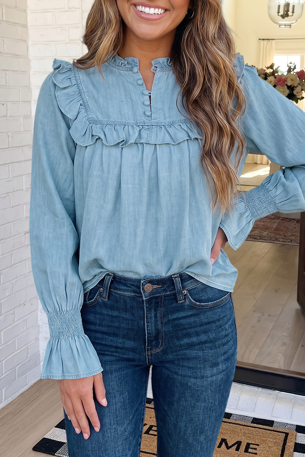 A woman wearing a light blue long-sleeve blouse with ruffle details and dark blue jeans, standing indoors in front of a white brick wall and mirror.