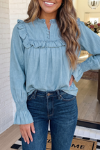 A woman wearing a light blue long-sleeve blouse with ruffle details and dark blue jeans, standing indoors in front of a white brick wall and mirror.