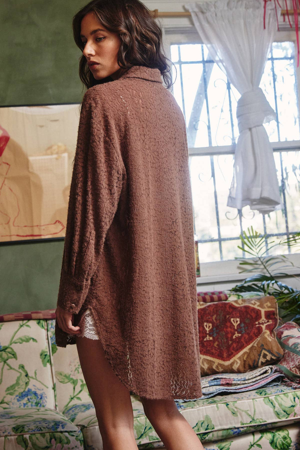 A woman wearing a brown textured knit cardigan sweater, photographed in a cozy, vintage-style room with floral wallpaper and a window in the background.