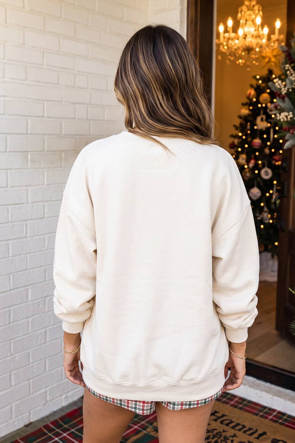 A woman viewed from behind wearing an oversized cream-colored crewneck sweatshirt, standing in front of a white brick wall with a decorated Christmas tree visible in the background.