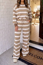 A woman models a matching two-piece lounge set featuring a long-sleeve sweatshirt and wide-leg pants, both with horizontal beige and off-white stripes. She is standing on a doormat in front of a white brick wall.