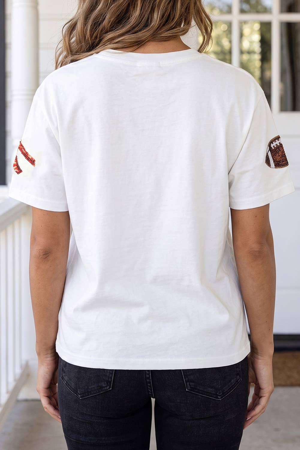 A woman viewed from behind wearing a white short-sleeve crewneck t-shirt with a distressed red stripe detail on the left sleeve and a football patch on the right sleeve, paired with dark jeans.
