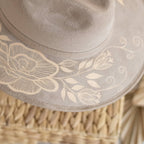 Close-up of a taupe felt hat with an artisan painted cream floral design  on the brim, resting on a woven straw surface. Crafted in Oaxaca, Mexico.