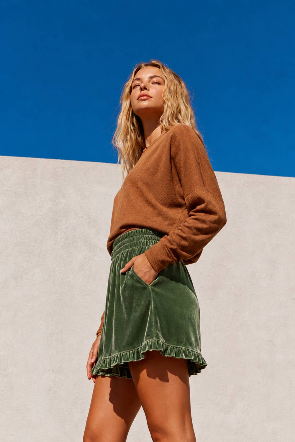 A woman wearing a brown knit sweater and an olive green velvet mini skirt with a ruffled hem, standing against a split background of blue sky and a light gray wall.