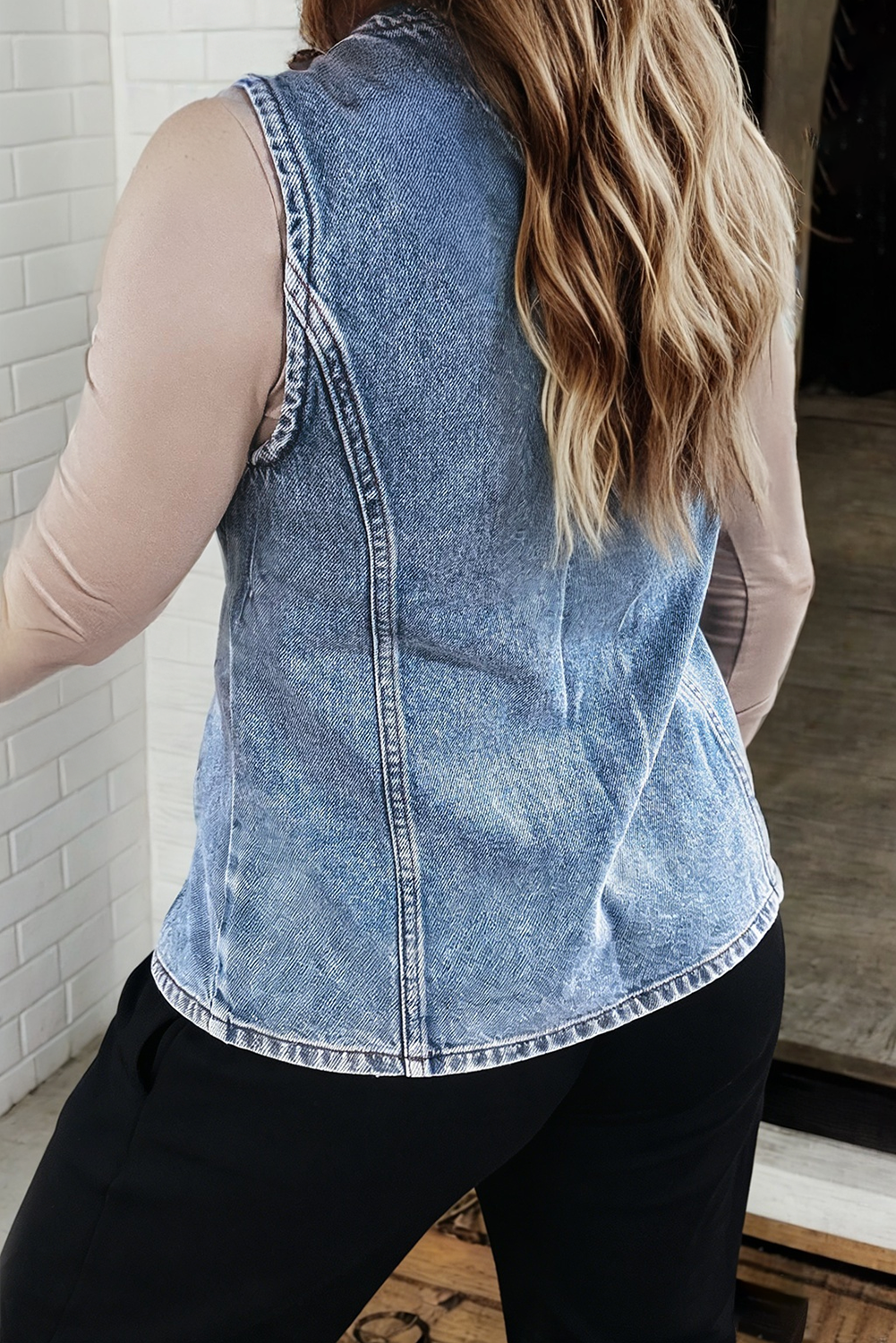 A woman shown from the back wearing a sleeveless light blue denim vest over a beige long-sleeve top and black pants, standing indoors near a white brick wall.