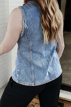A woman shown from the back wearing a sleeveless light blue denim vest over a beige long-sleeve top and black pants, standing indoors near a white brick wall.