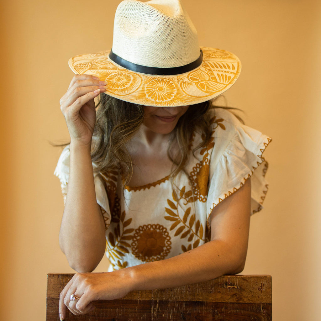 Woman wearing a decorative artisan painted sun hat and patterned top against a warm background. Crafted in Oaxaca, Mexico.