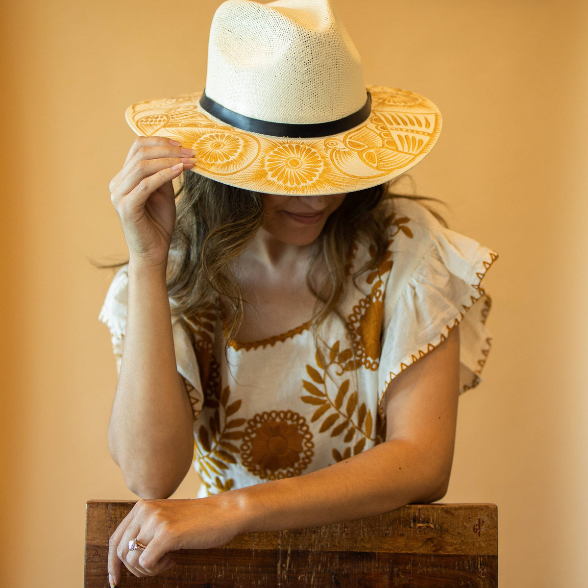 Woman wearing a decorative artisan painted sun hat and patterned top against a warm background. Crafted in Oaxaca, Mexico.