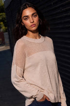 A woman wearing a light beige knit sweater with a sheer mesh yoke, photographed outdoors against a dark metal shutter.