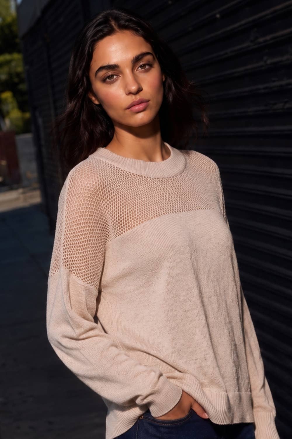 A woman wearing a light beige knit sweater with a sheer mesh yoke, photographed outdoors against a dark metal shutter.