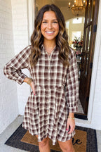 A woman smiling while wearing a brown and beige plaid button-down shirt dress, standing on a porch in front of a white brick wall and open doorway.