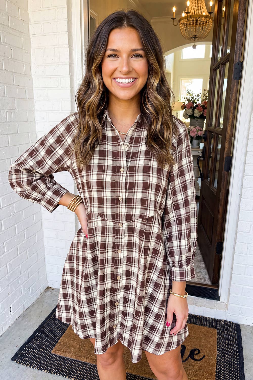 A woman smiling while wearing a brown and beige plaid button-down shirt dress, standing on a porch in front of a white brick wall and open doorway.