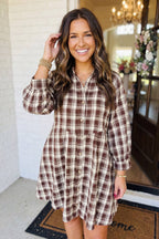 A woman wearing a brown and beige plaid button-down shirt dress, standing in front of a white brick wall and a doorway.