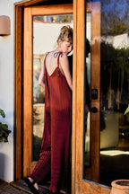 A woman wearing a rust-brown pleated maxi dress with tie-shoulder straps and a backless open back, standing in a doorway of a wooden door frame.