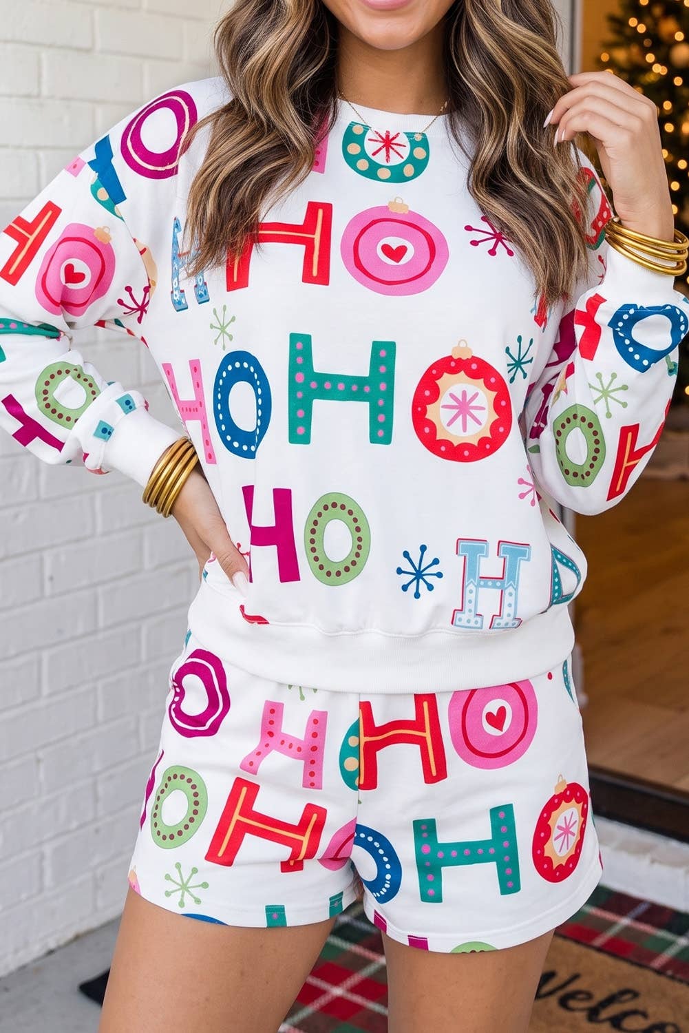A woman wearing a matching white holiday sweatshirt and shorts set with a colorful 'HO HO HO' print, standing in front of a white brick wall.