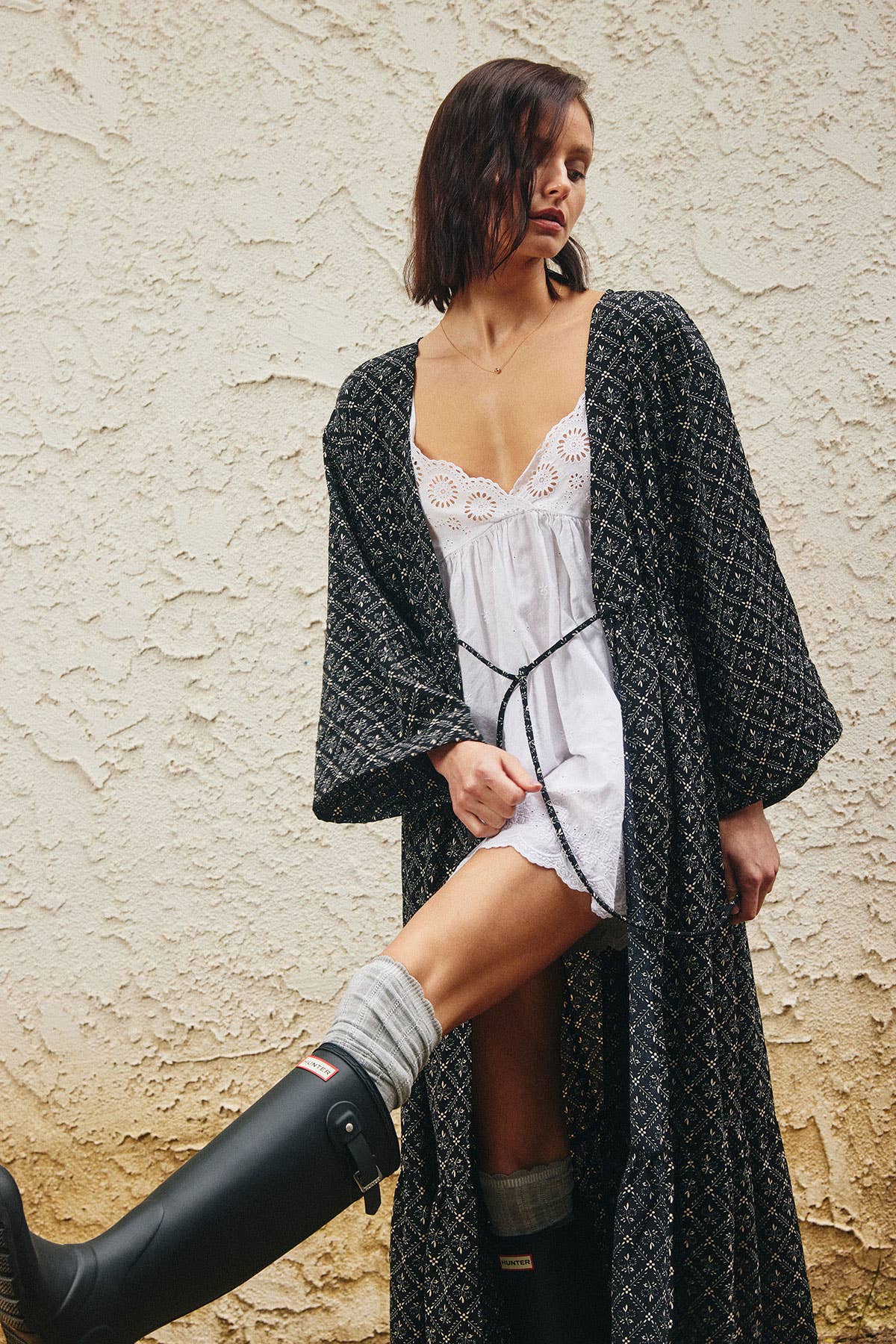 A woman stands against a textured beige wall wearing a long black patterned robe over a white lace-trim nightgown, with gray socks and black rain boots.