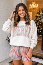 A woman wearing a cream-colored crewneck sweatshirt with 'MERRY' in red plaid letters, paired with matching red and green plaid shorts, standing in a decorated holiday home.
