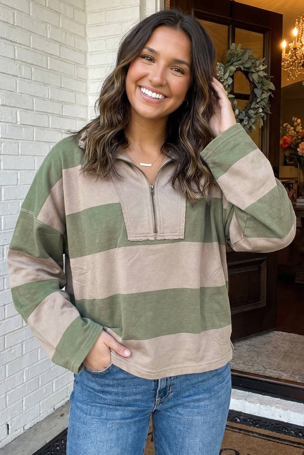 A woman smiling and posing in a long-sleeve quarter-zip pullover sweater with horizontal stripes in sage green and beige, paired with blue jeans.
