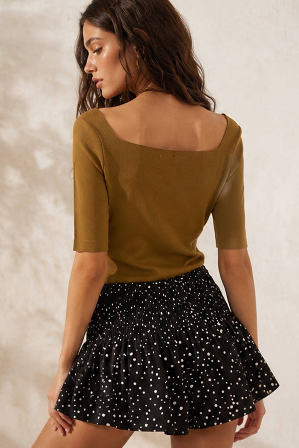 A woman shown from the back wearing a mustard yellow short-sleeve top with a wide scoop neckline and a black flared skirt with white polka dots, set against a textured light beige wall.