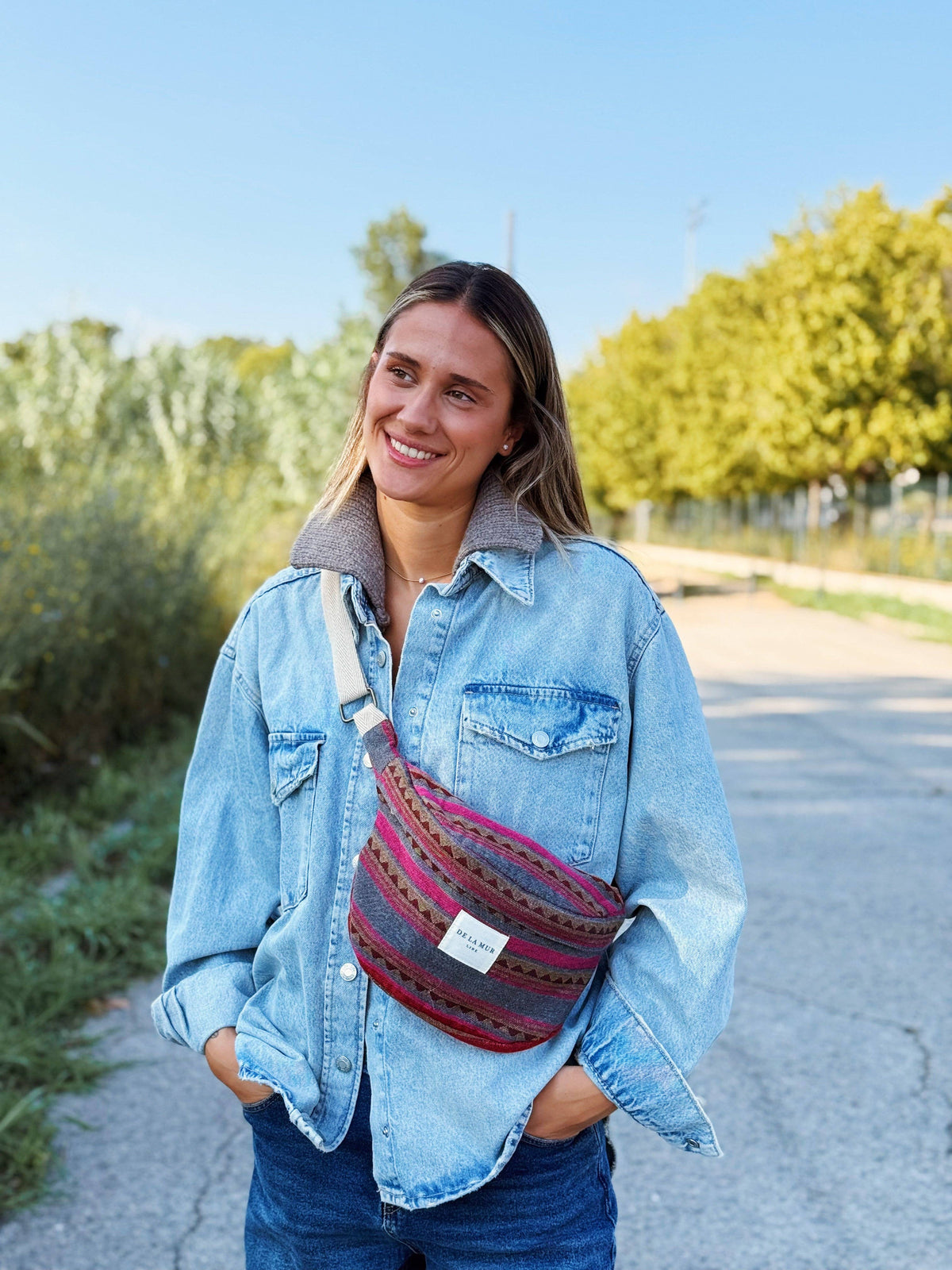 Smiling woman in a denim jacket wearing the pink and teal striped jacquard fanny pack crossbody outdoors