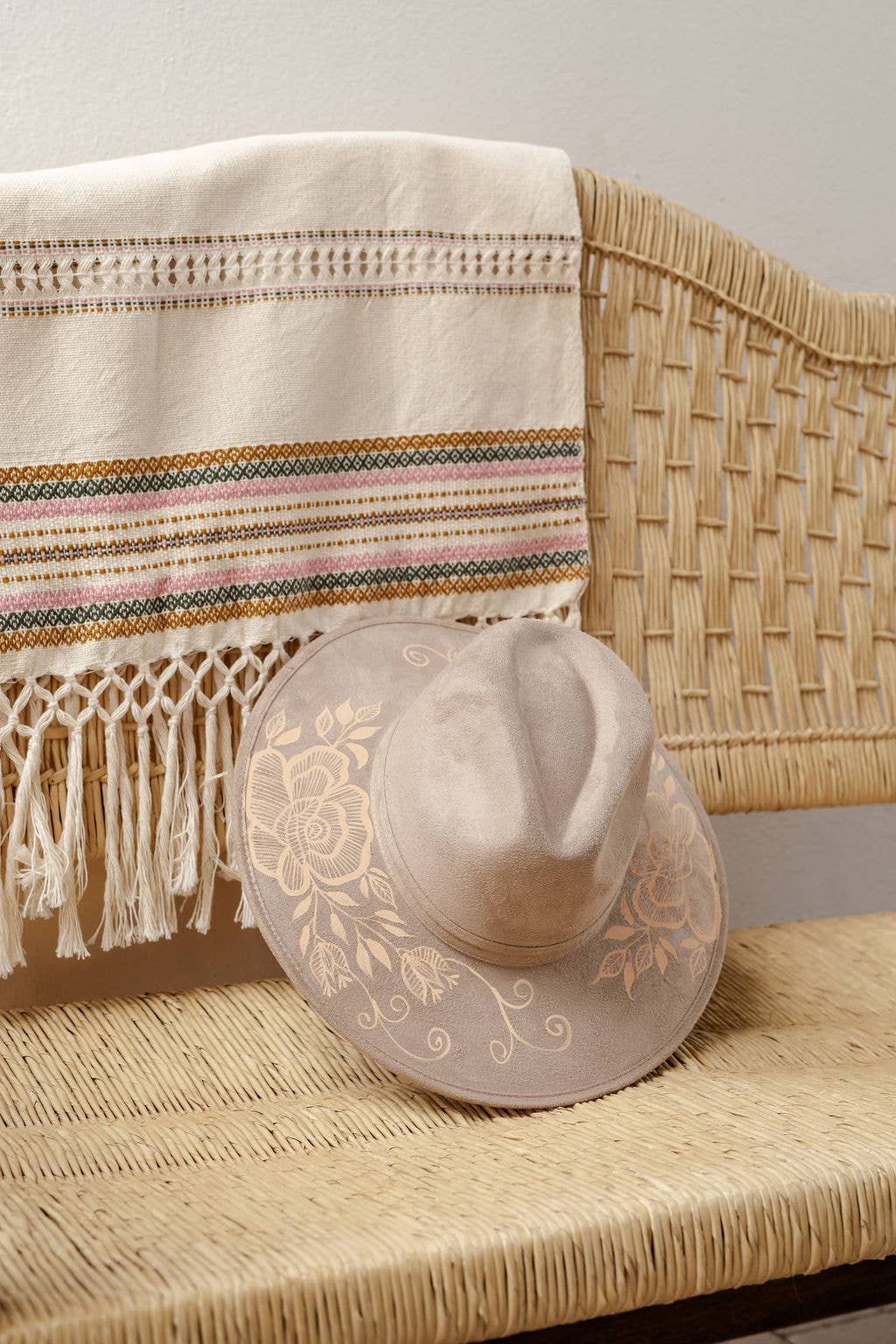 A light beige wide-brimmed hat with an artisan painted floral design rests on a woven rattan bench next to a cream-colored Mexican-style woven blanket with colorful stripes and fringe. Crafted in Oaxaca, Mexico.