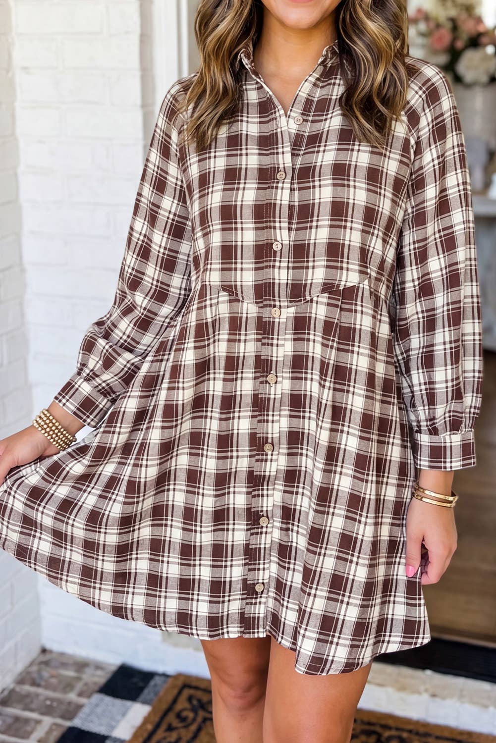 A woman wearing a brown and beige plaid button-up shirt dress, standing in front of a white brick wall.