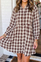 A woman wearing a brown and beige plaid button-up shirt dress, standing in front of a white brick wall.