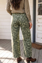A person standing on a wooden porch wearing wide-leg sage green pants with a beige paisley pattern, paired with a tan long-sleeve shirt and brown ankle boots.