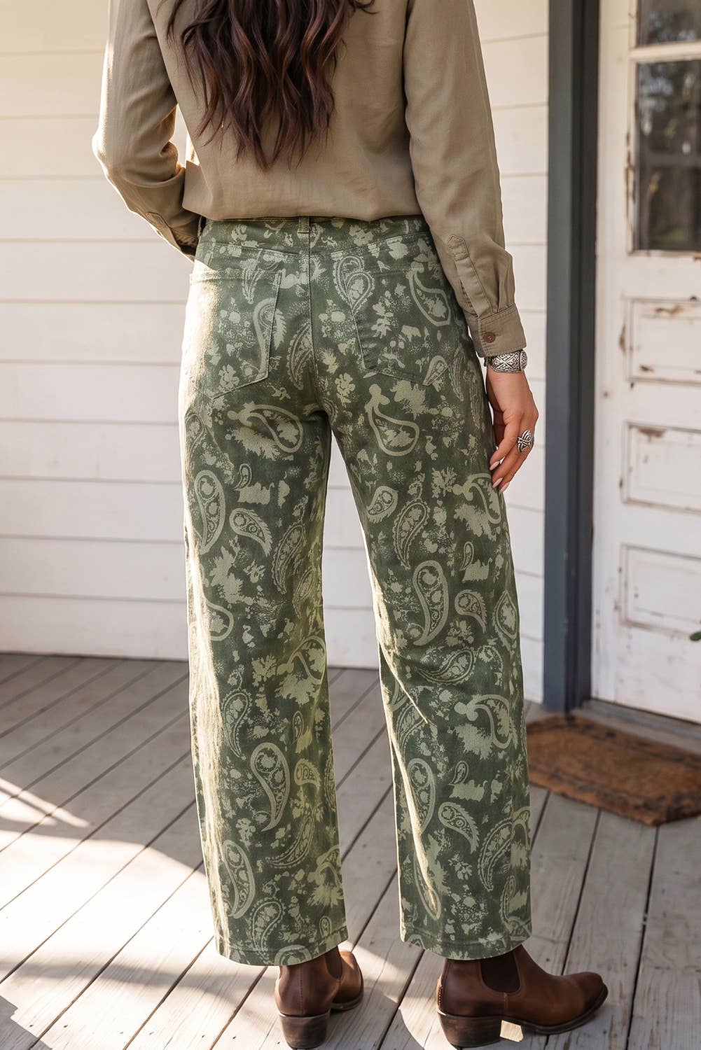A person standing on a wooden porch wearing wide-leg sage green pants with a beige paisley pattern, paired with a tan long-sleeve shirt and brown ankle boots.