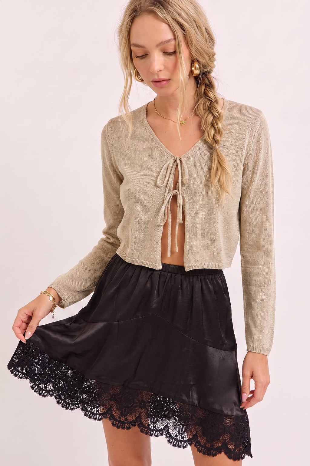 A woman modeling a beige cropped knit cardigan with a tie-front detail and a black satin mini skirt with a scalloped lace hem, against a plain light background.