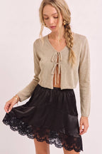 A woman modeling a beige cropped knit cardigan with a tie-front detail and a black satin mini skirt with a scalloped lace hem, against a plain light background.
