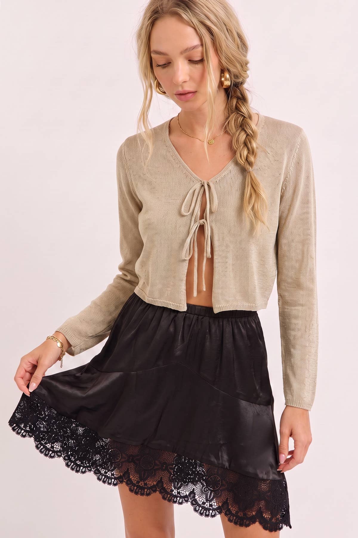 A woman modeling a beige cropped knit cardigan with a tie-front detail and a black satin mini skirt with a scalloped lace hem, against a plain light background.