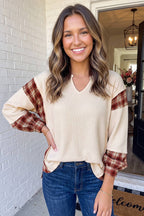 A smiling woman wearing a cream waffle-knit V-neck top with rust red and brown plaid balloon sleeves, paired with blue jeans, standing in front of a white brick wall doorway.
