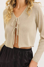 A woman wearing a beige knit cropped cardigan with a V-neck and tie-front detail, paired with black bottoms, against a plain light background.