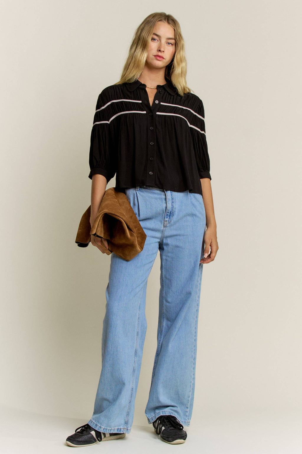 A woman modeling a black button-up blouse with white horizontal stripes on the chest, paired with light blue wide-leg jeans, black sneakers, and holding a brown suede jacket against a plain beige studio background.
