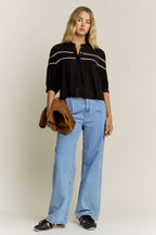 A woman modeling a black button-up blouse with white horizontal stripes on the chest, paired with light blue wide-leg jeans, black sneakers, and holding a brown suede jacket against a plain beige studio background.