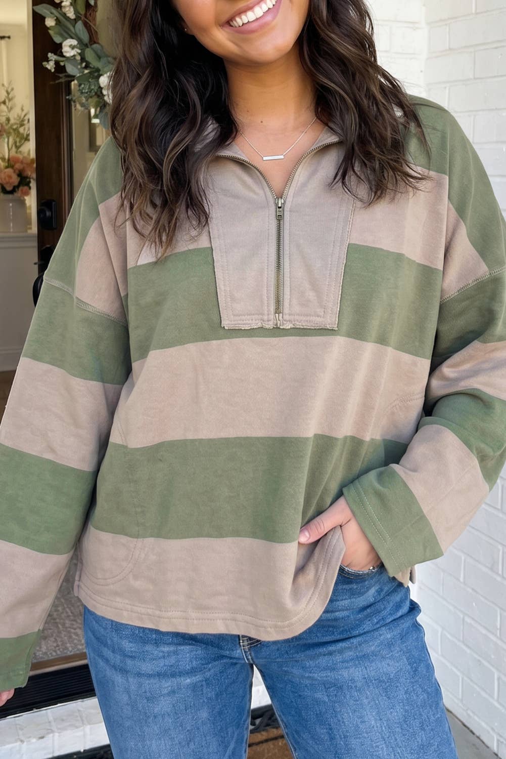 A woman wearing a long-sleeve quarter-zip pullover top with horizontal stripes in sage green and beige, paired with blue jeans.