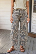 A person wearing wide-leg cropped jeans with a muted camouflage pattern in gray, beige, and brown, paired with a light beige top and brown slide sandals, standing on a wooden deck.