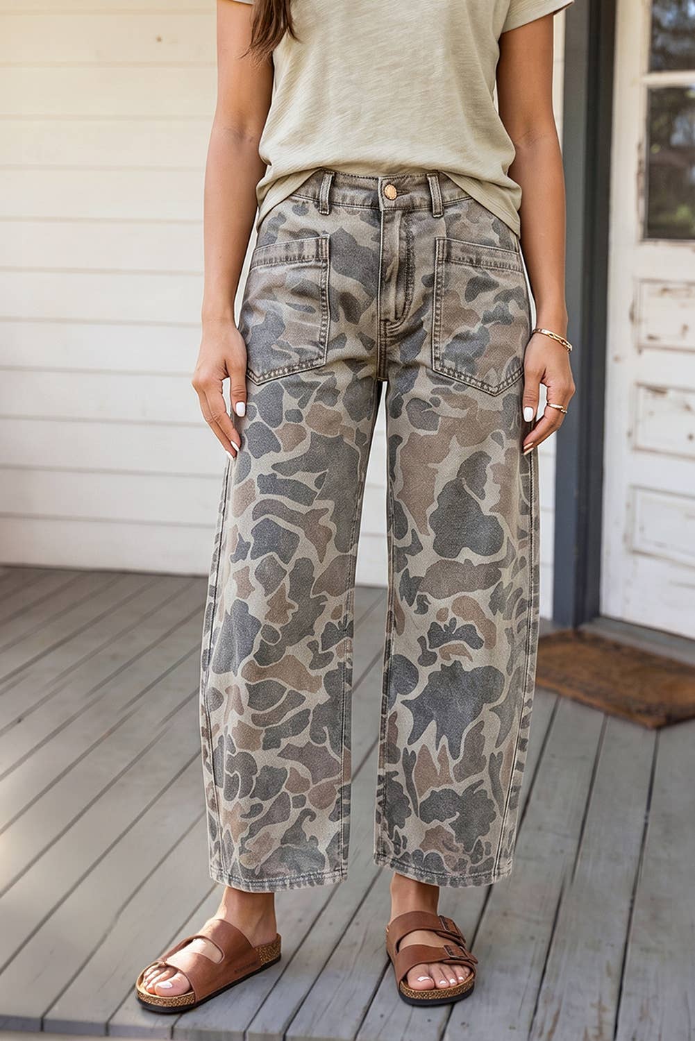 A person wearing wide-leg cropped jeans with a muted camouflage pattern in gray, beige, and brown, paired with a light beige top and brown slide sandals, standing on a wooden deck.