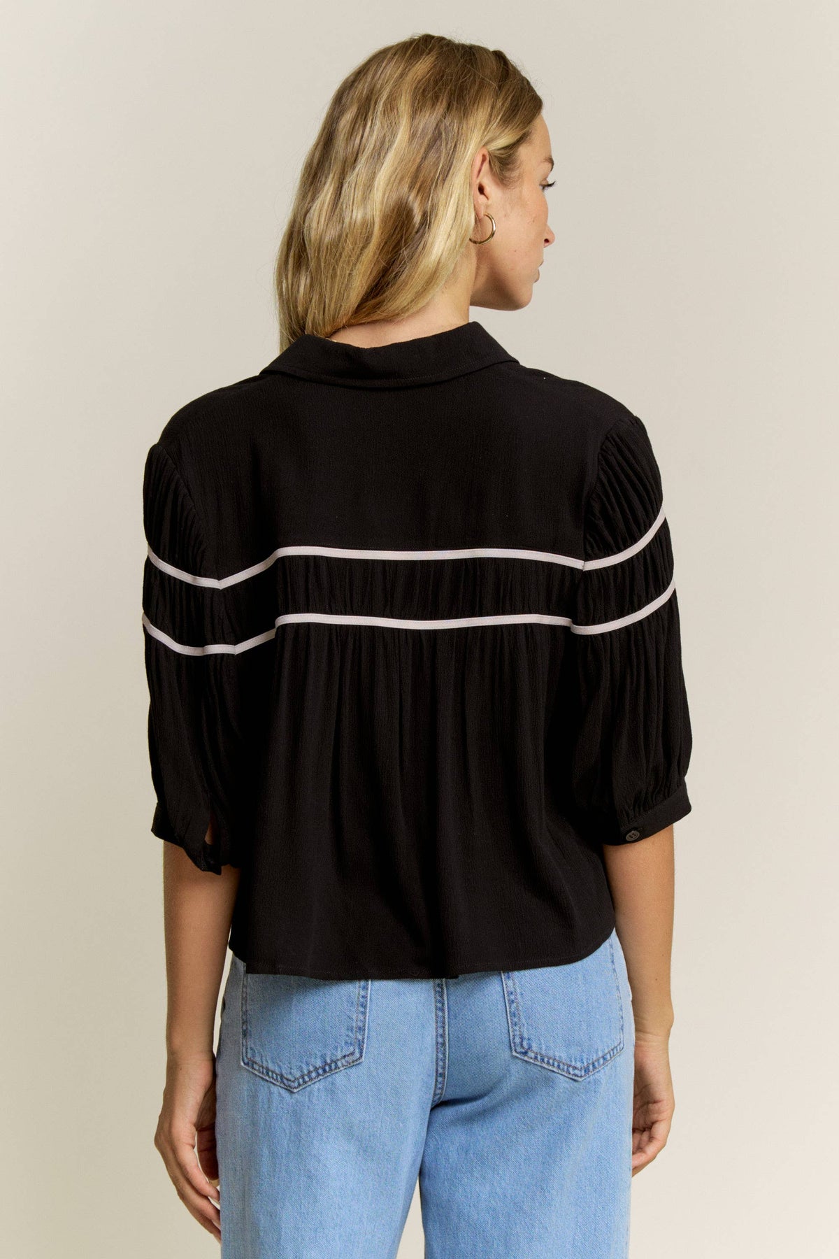 A woman viewed from the back wearing a black blouse with horizontal white stripe details across the shoulders and upper back, paired with light blue jeans, against a plain light beige studio background.
