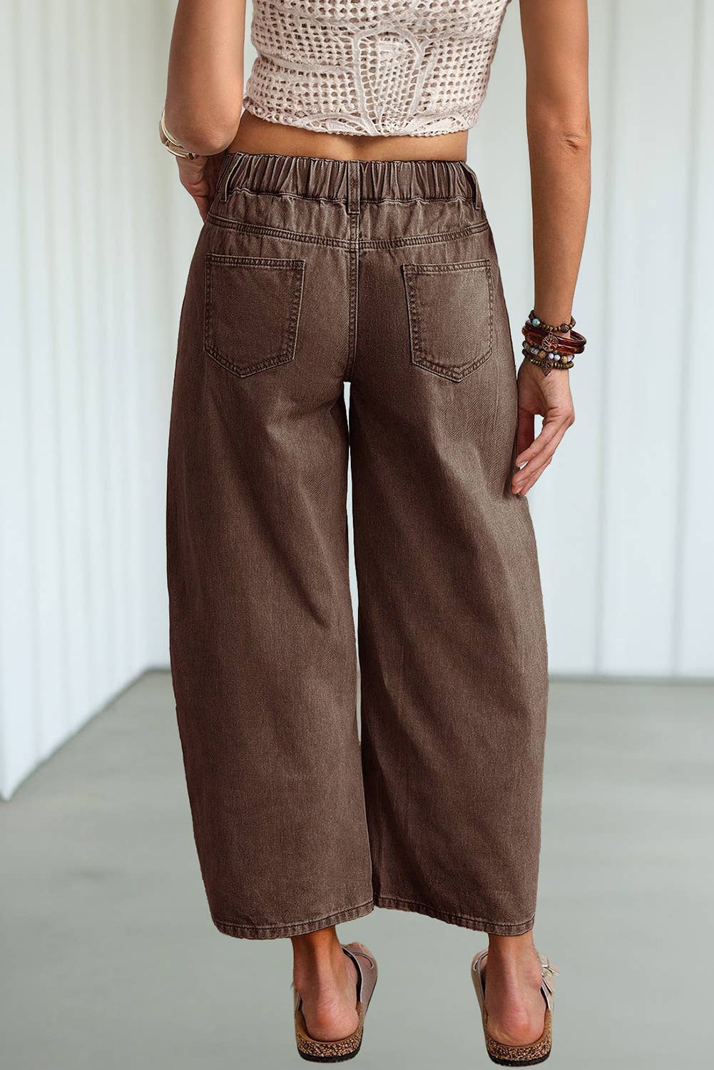 A person shown from the back wearing loose, wide-leg cropped pants in a medium brown color, paired with a cream crochet crop top and brown wedge sandals.