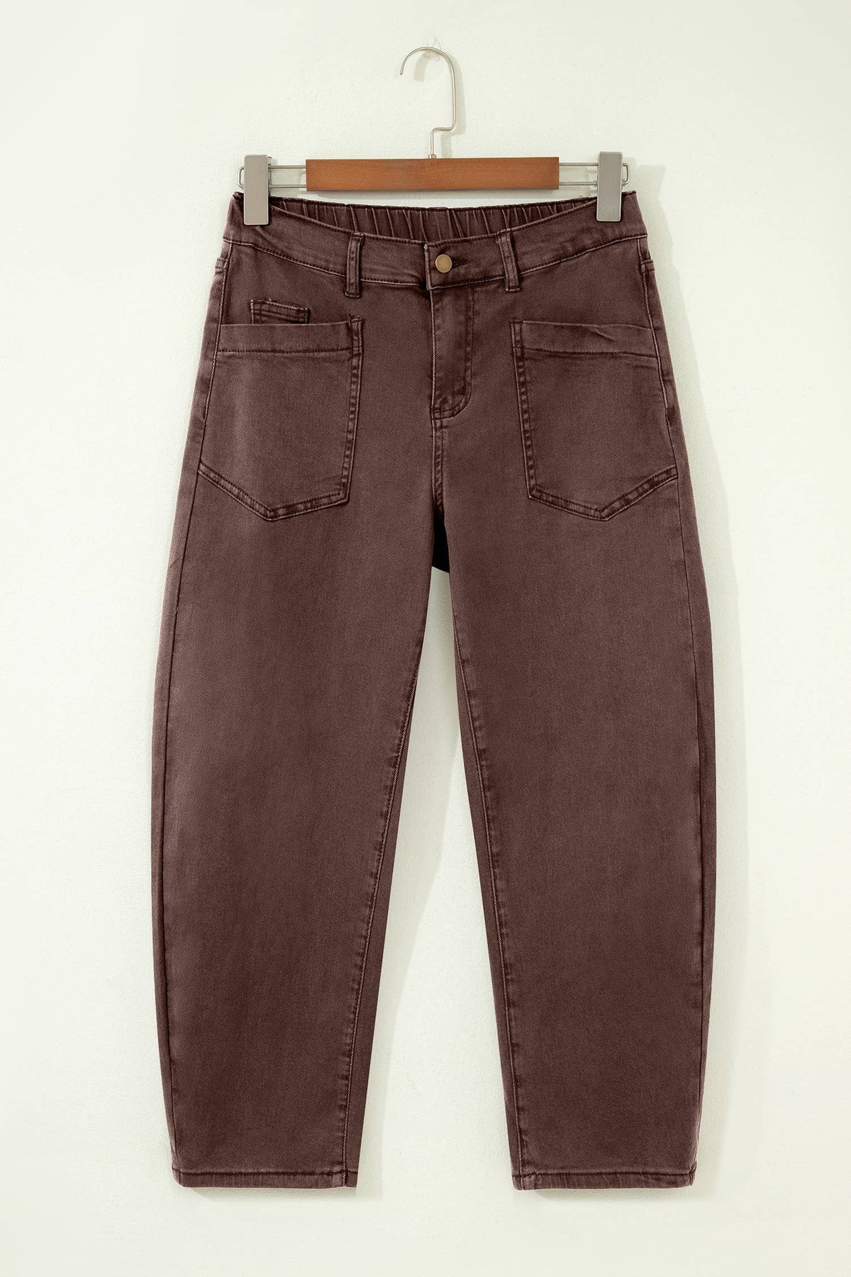 A pair of dark brown washed denim jeans hanging on a wooden hanger against a plain white wall.