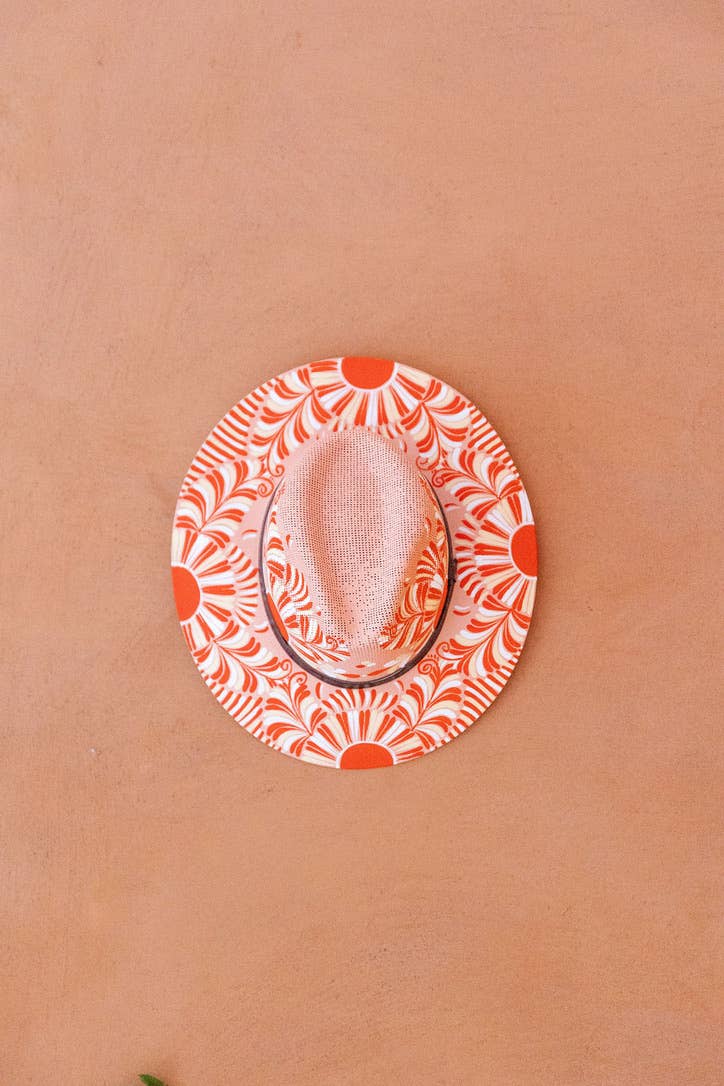A fedora-style hat with a white crown and brim featuring an artisan painted vibrant orange sunburst and leaf pattern, resting on a textured terracotta surface. Crafted in Oaxaca, Mexico.