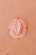 A fedora-style hat with a white crown and brim featuring an artisan painted vibrant orange sunburst and leaf pattern, resting on a textured terracotta surface. Crafted in Oaxaca, Mexico.