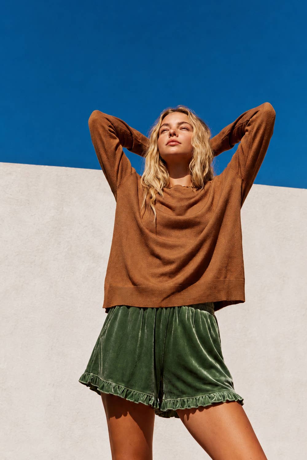 A woman posing outdoors against a split background of blue sky and a light beige wall, wearing a brown long-sleeve knit top and green velvet shorts.