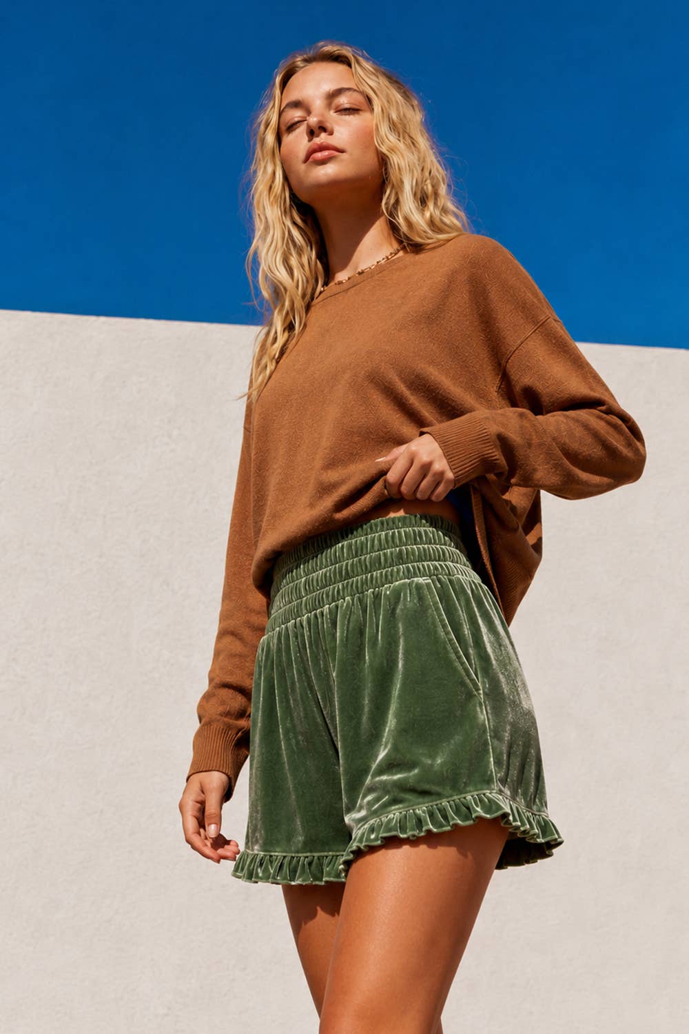 A woman wearing a brown long-sleeve knit sweater and green velvet shorts with a ruffled hem, standing against a split blue and white background.